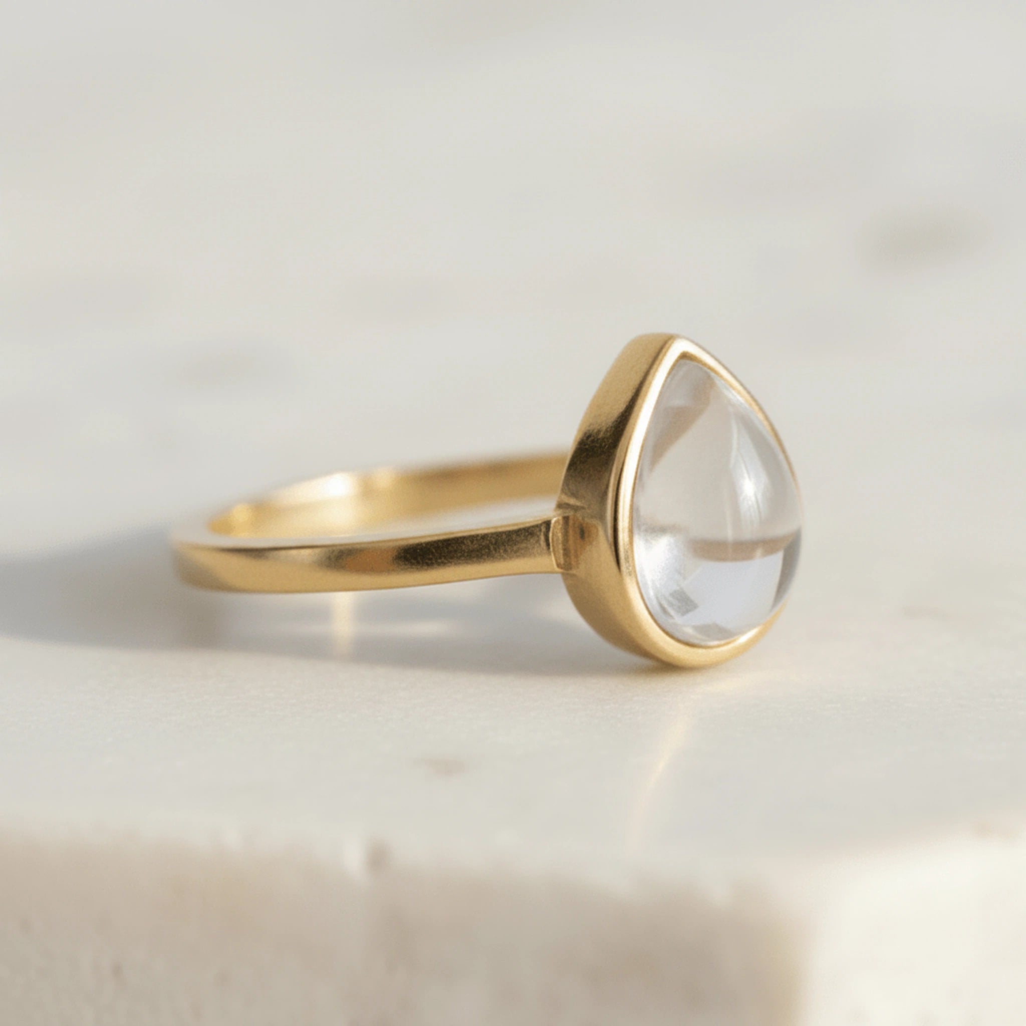 Gold April Birthstone Ring with a teardrop-shaped cabochon and bezel set natural clear quartz gemstone on a light background