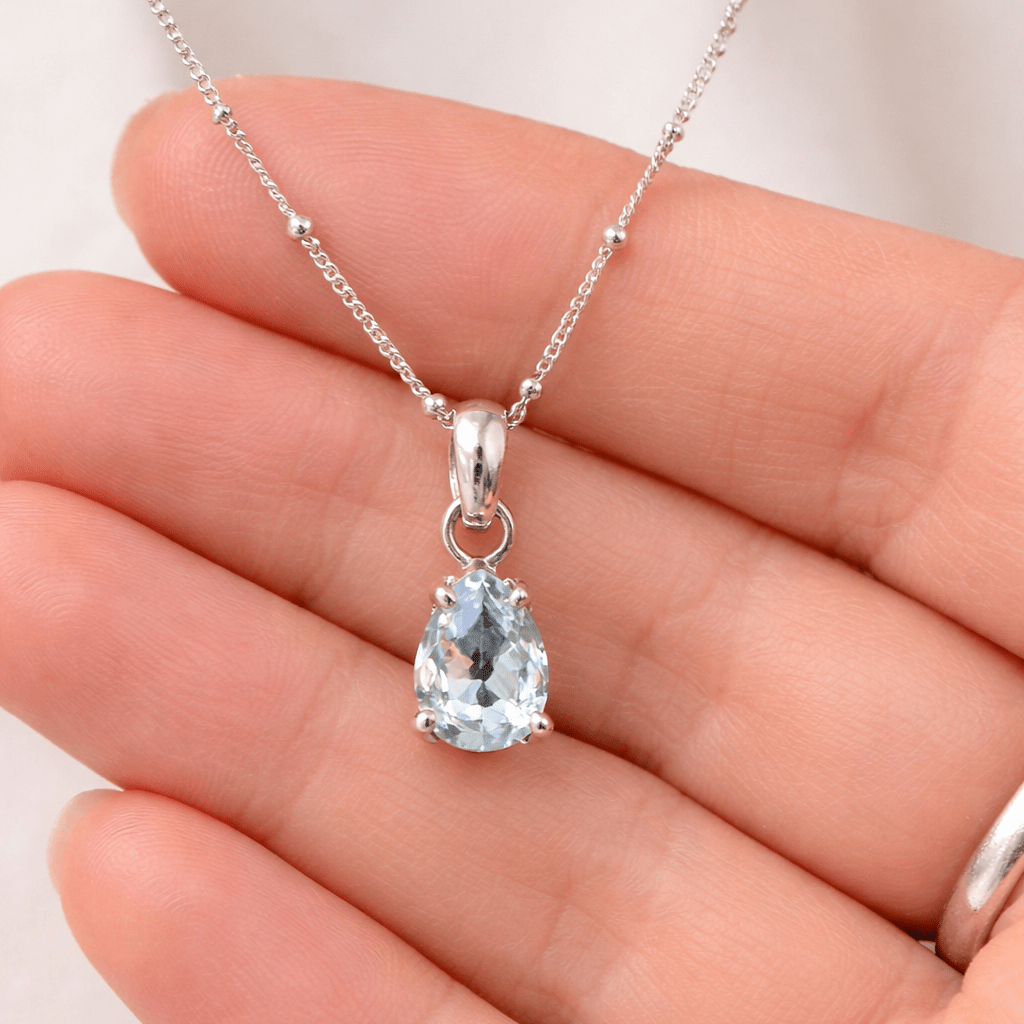 Silver necklace with a teardrop-shaped blue aquamarine gemstone held in a hand against a white background