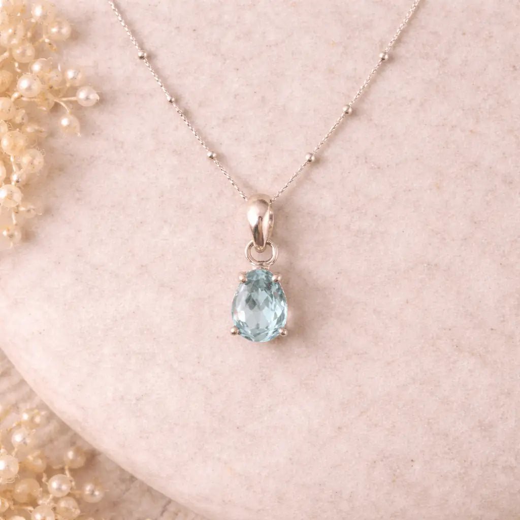 Necklace with a teardrop-shaped aquamarine faceted claw set blue gemstone pendant on a soft pink background