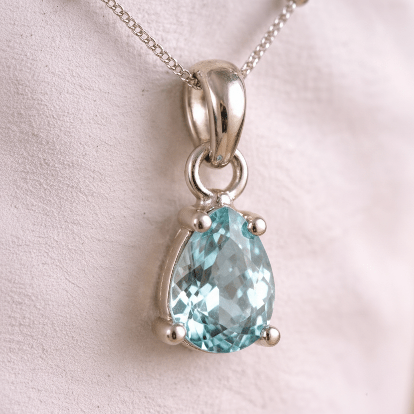 Silver pendant with a teardrop-shaped blue aquamarine gemstone on a light pink background