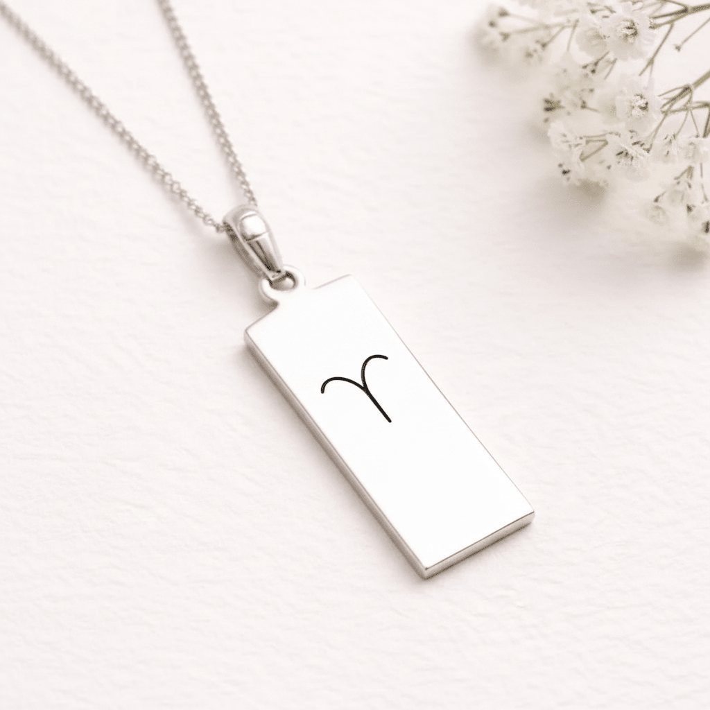A silver rectangular pendant necklace with a black engraved zodiac symbol, displayed on a textured white surface with small white flowers in the corner.