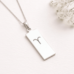A silver rectangular pendant necklace with a black engraved zodiac symbol, displayed on a textured white surface with small white flowers in the corner.