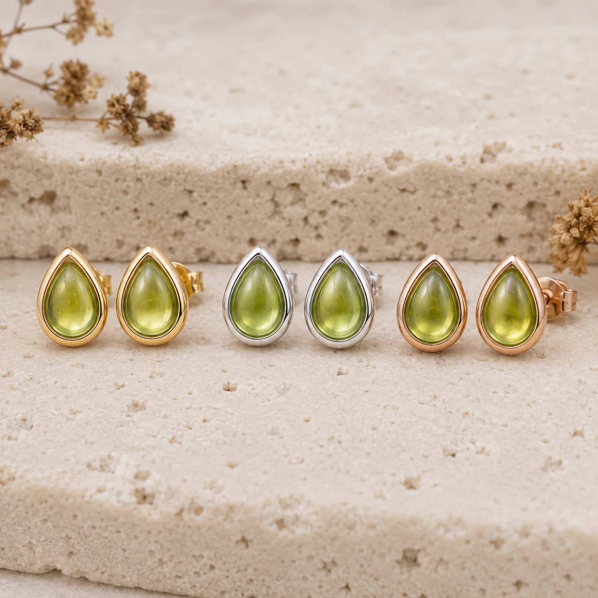 Three pairs of August Birthstone Natural Peridot Earrings - Six teardrop-shaped stud earrings with olive green gemstones in gold, rose gold, and silver settings, arranged in a row on a textured beige stone surface with dried flowers.