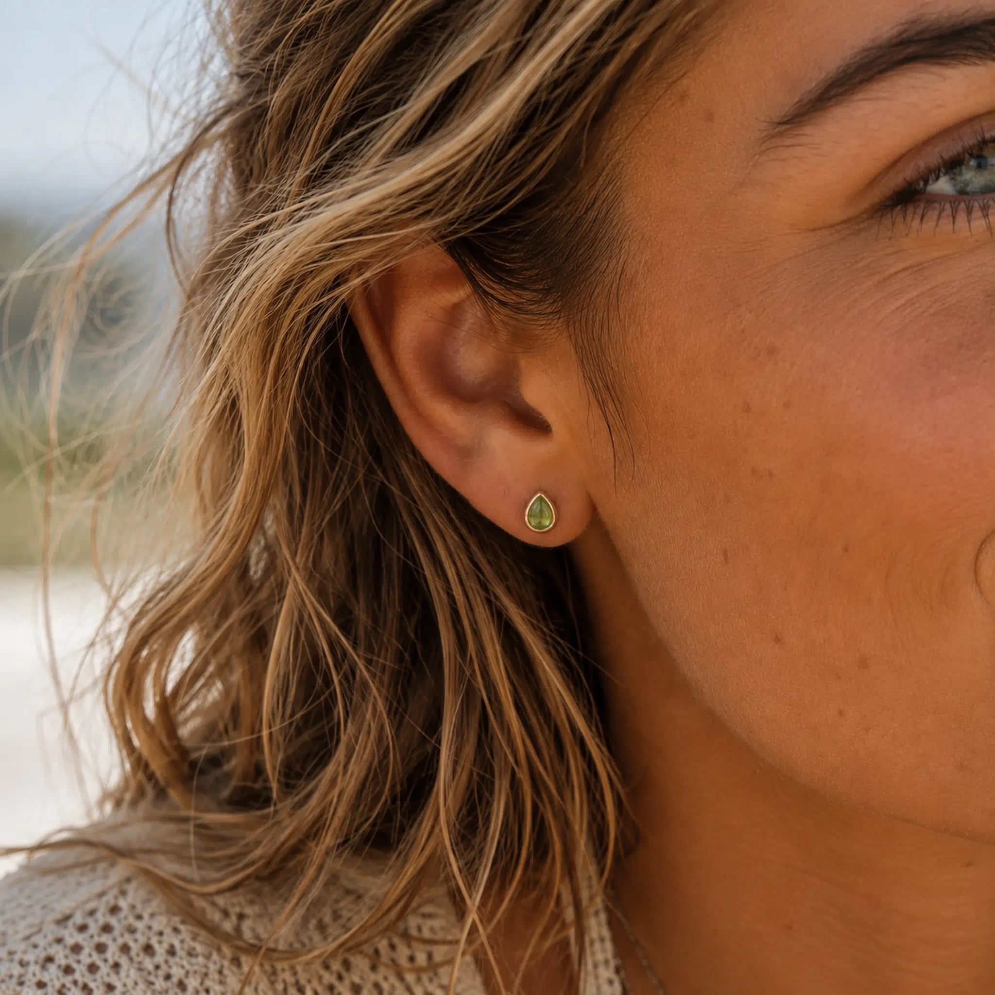 A close-up of a woman's ear wearing August Birthstone Natural Peridot Earrings - small teardrop-shaped green stud earring with a gold-tone outline.