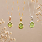 Three August Birthstone Necklaces featuring teardrop pendant necklaces with light green natural peridot gemstone pendants in rose gold, yellow gold, and silver settings, hanging against a warm beige background with delicate dried flowers.