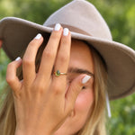 A close-up lifestyle shot of a woman wearing a wide-brimmed taupe hat and white nail polish, holding her hand up to showcase a delicate gold ring with a small green teardrop gemstone.