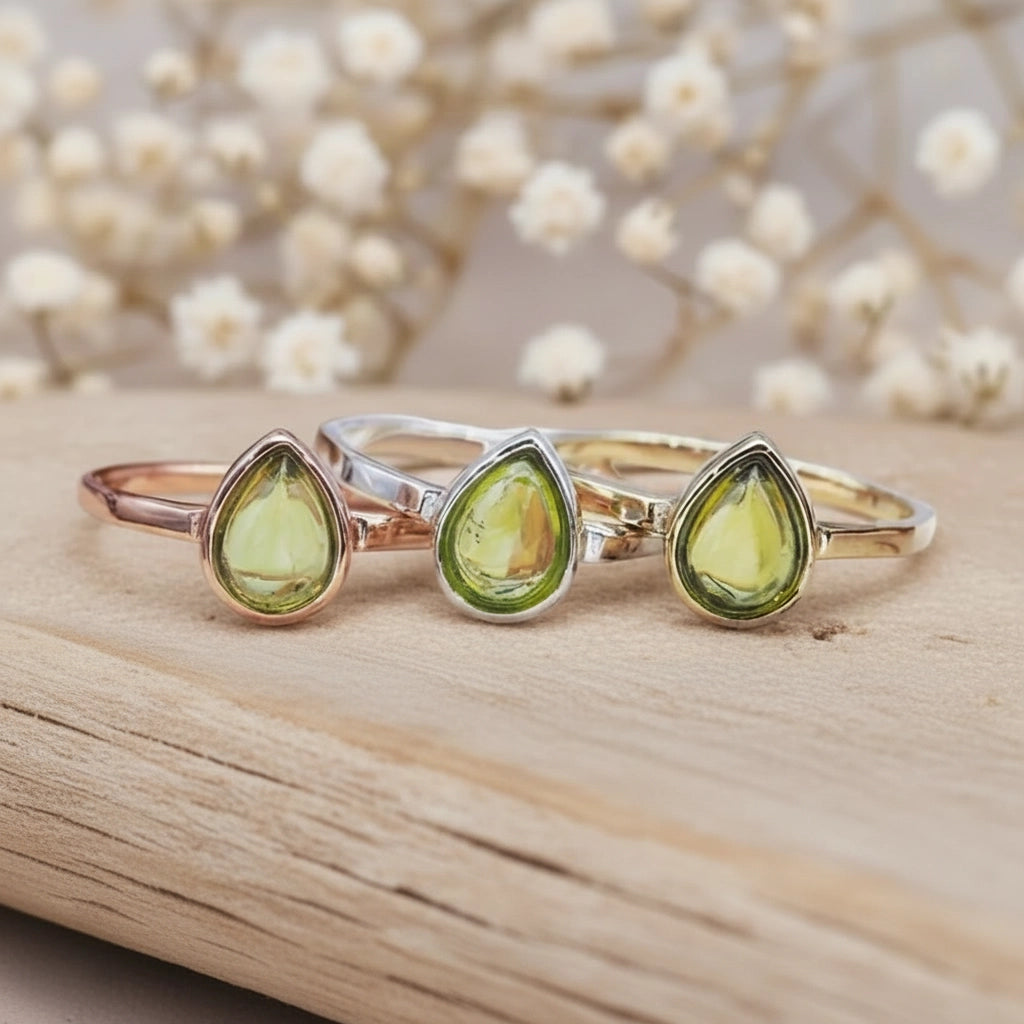 Three rings in Rose Gold, Silver and Gold with teardrop-shaped peridot gemstones on a wooden surface with a blurred floral background.