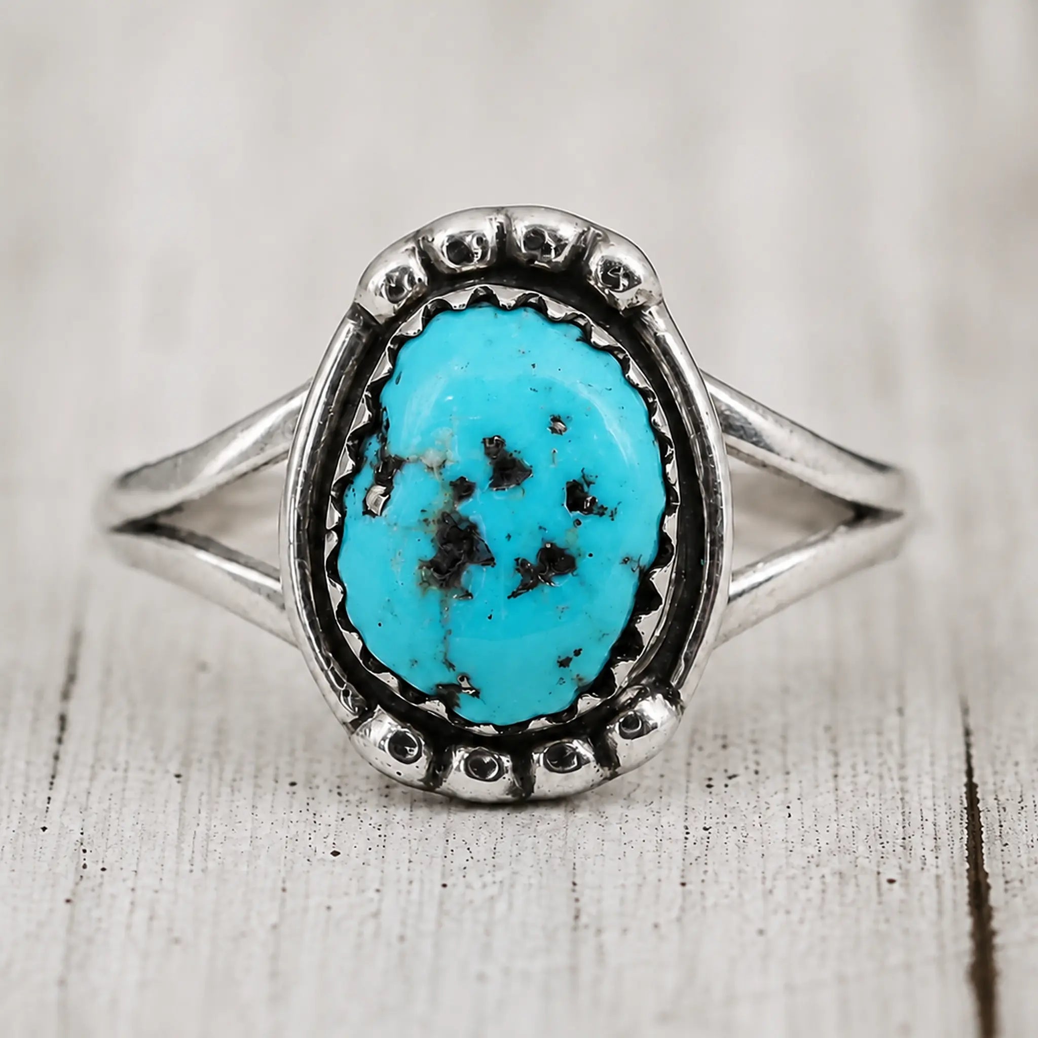 Front view of a sterling silver turquoise ring with an oval matrix stone and split band on pale whitewashed wood
