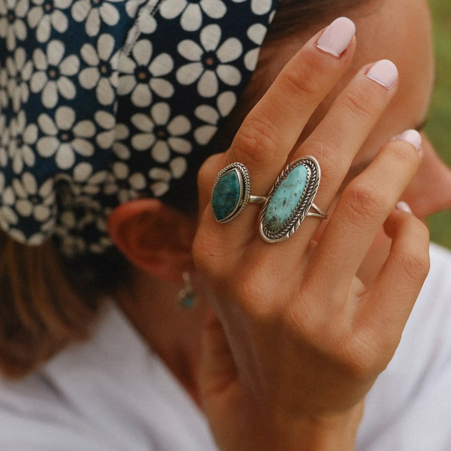 woman wearing a blue bandana with white daisies holding her hand in front of her face which has a turquoise ring and an azurite ring on it