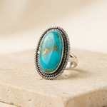 Front three-quarter view of a handmade Mohave composite turquoise ring with bright blue oval cabochon, metallic matrix and oxidised sterling silver twisted rope border