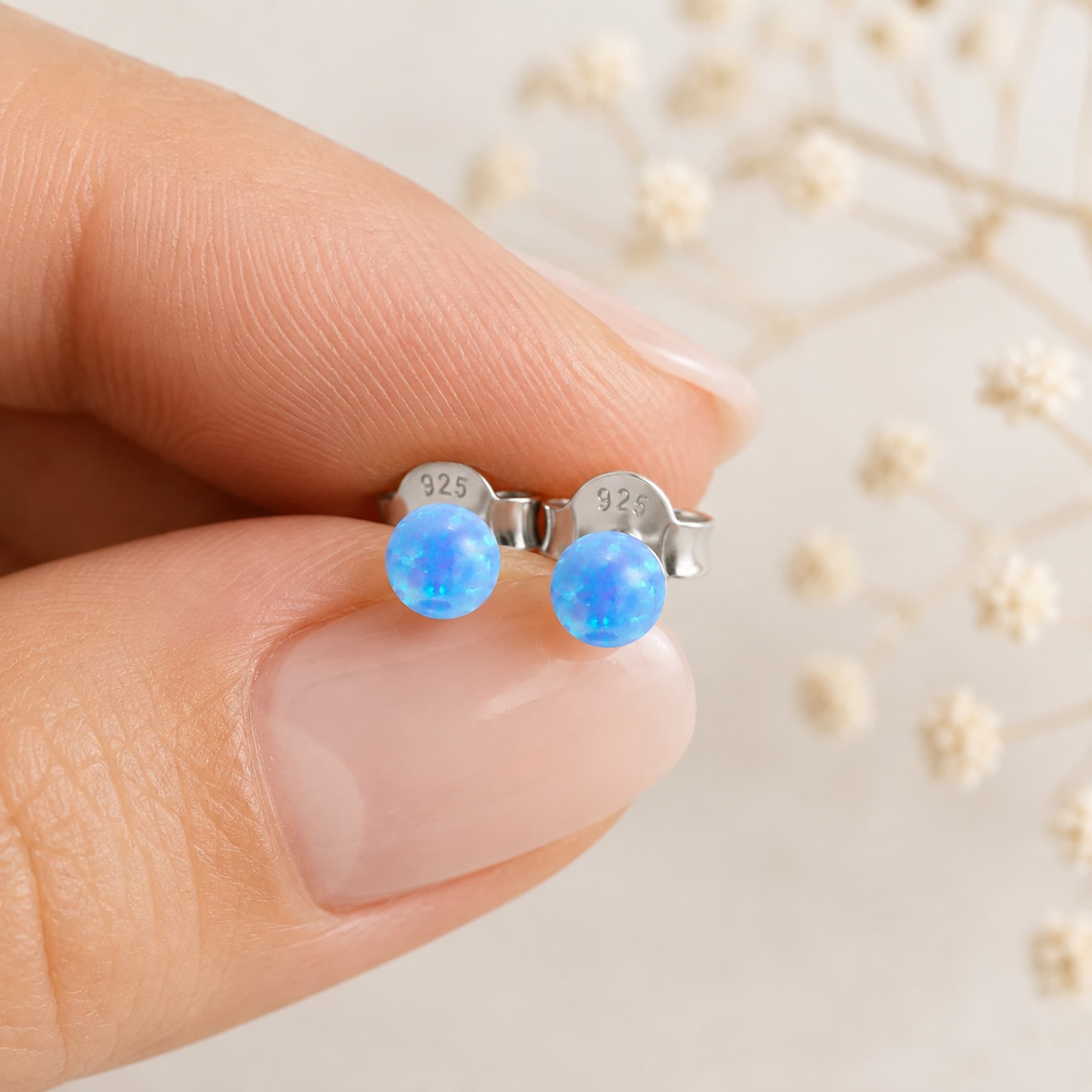 A close-up of a hand holding a pair of small silver stud earrings with round blue iridescent opal balls. The back of the earrings shows a stamped '925' mark.
