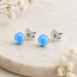 A pair of silver stud earrings with round, light blue opal balls, displayed on a light beige stone surface with dried white flowers.