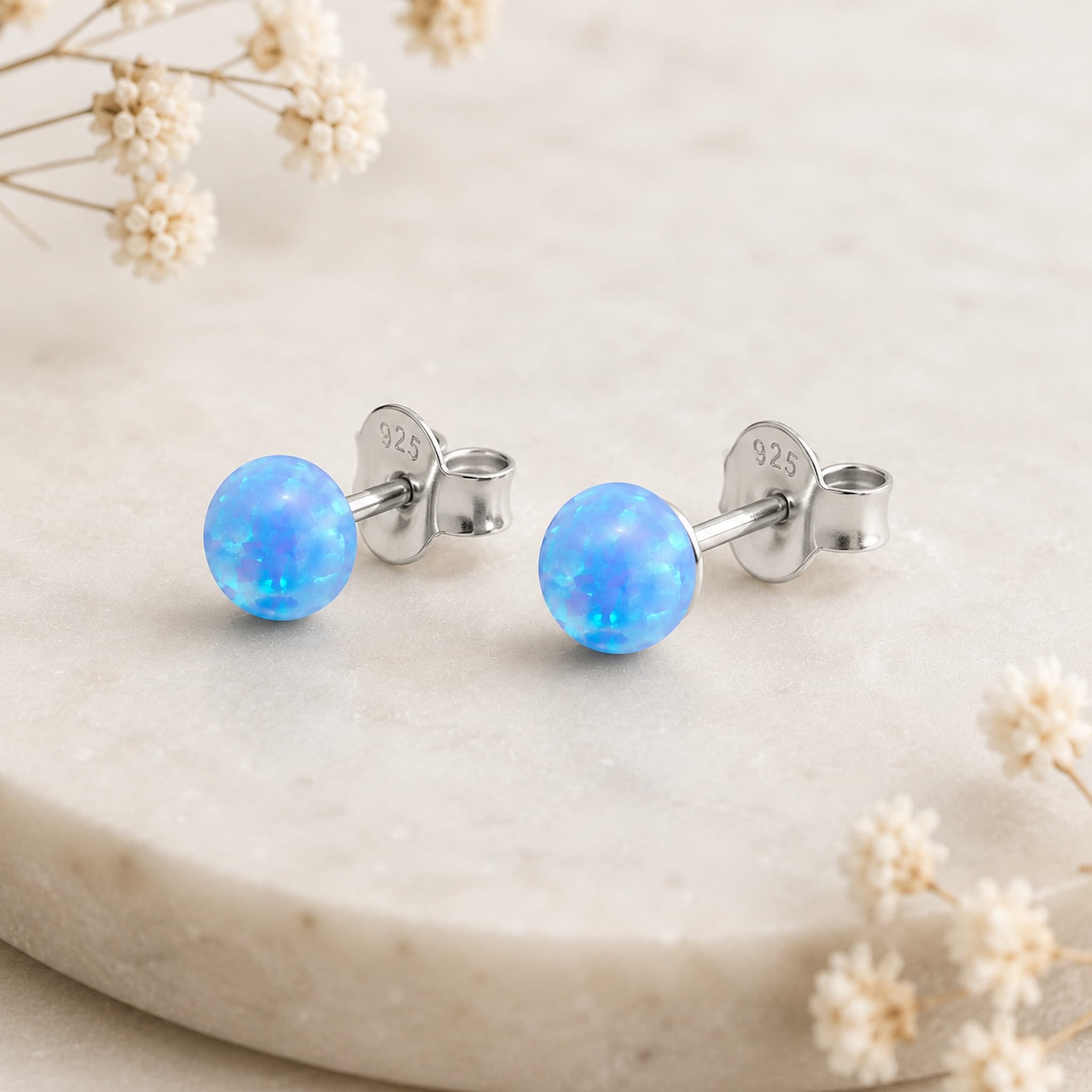 A pair of silver stud earrings with round, light blue opal balls, displayed on a light beige stone surface with dried white flowers.