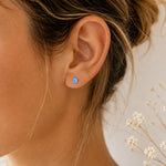 A close-up of a person's ear wearing a small round light blue opal stud earring, with light brown hair tied back and delicate white flowers in the background.