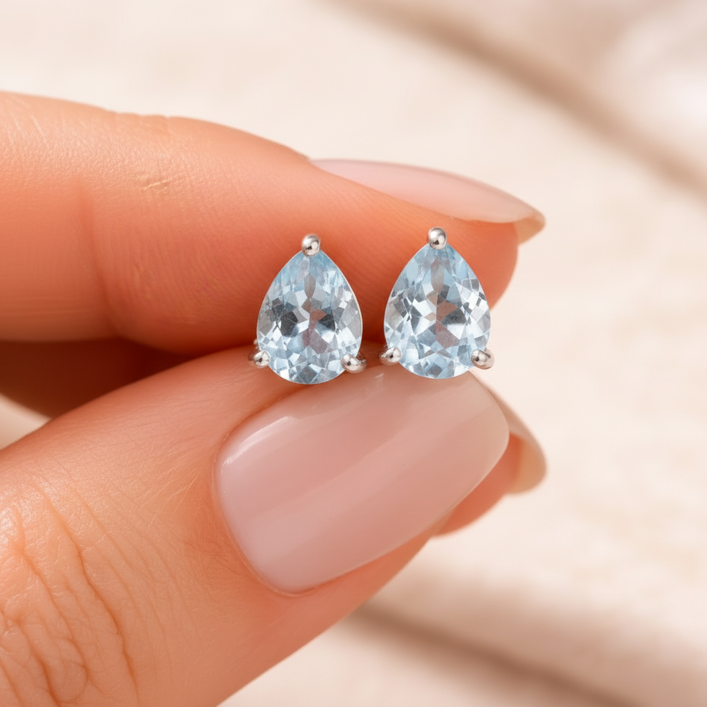 Teardrop-shaped blue topaz gemstone earrings held between fingers against a neutral background