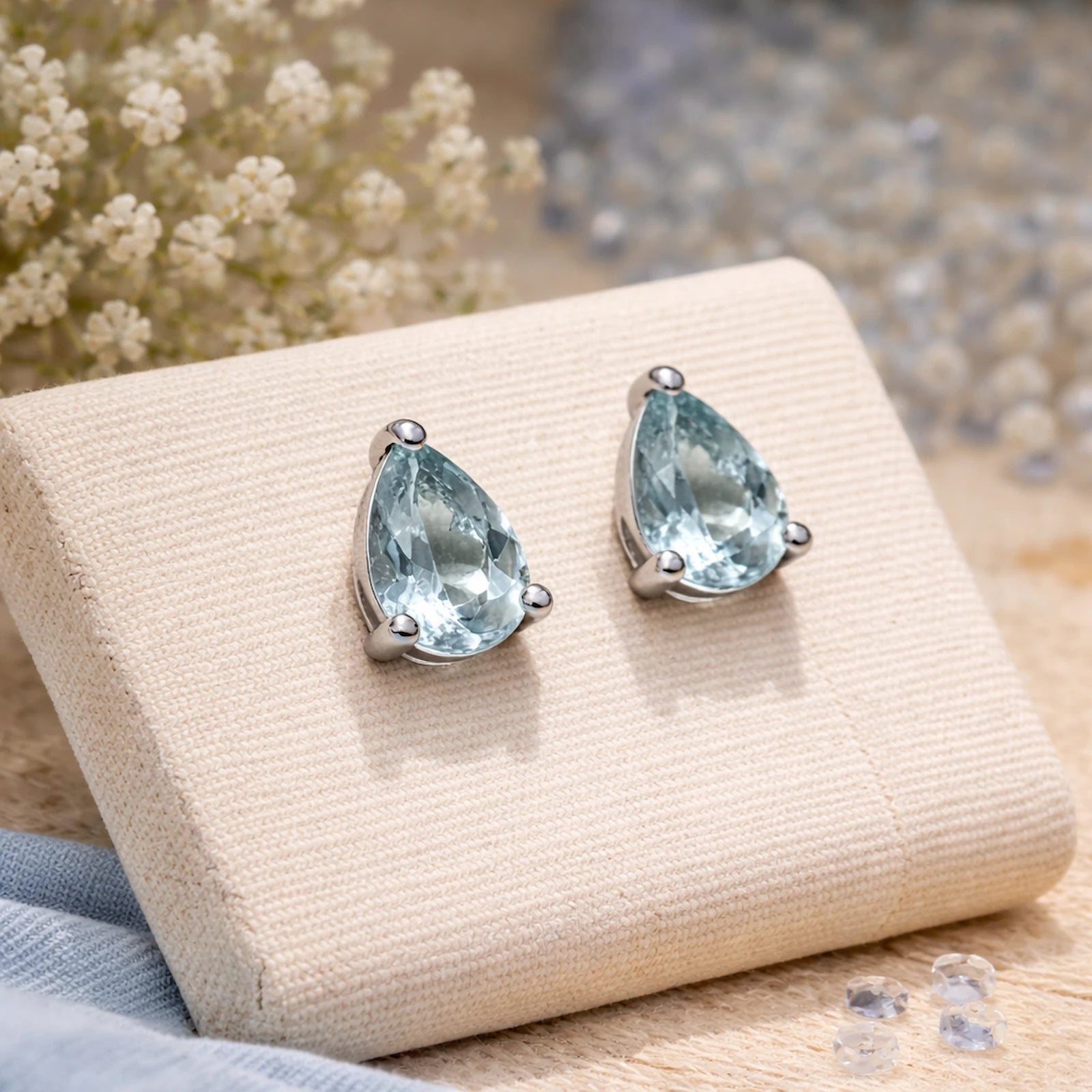 Teardrop-shaped blue gemstone earrings on a beige jewelry display with flowers in the background.