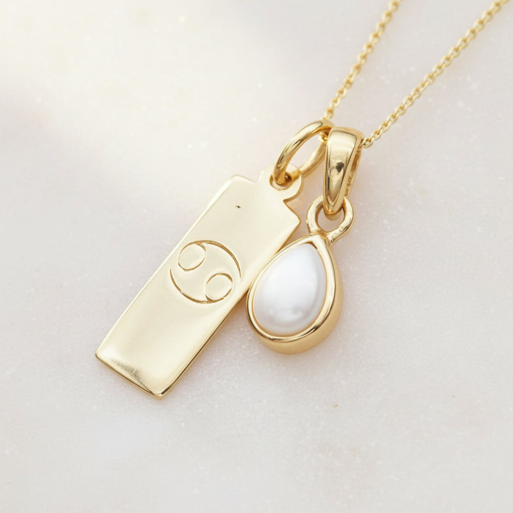 Gold necklace with a cancer zodiac pendant and pear shaped natural pearl pendant on a textured gold and gray background