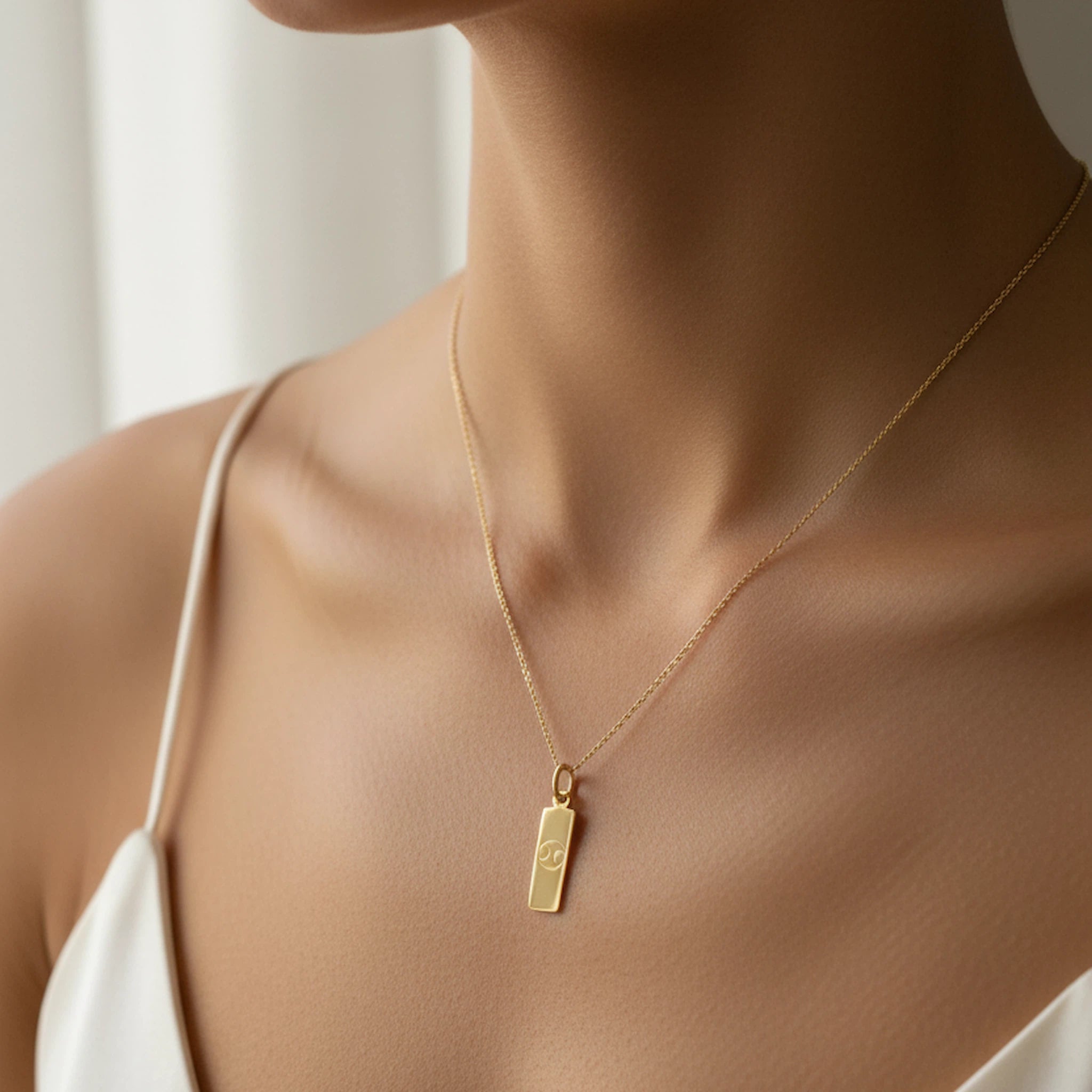 Dainty Gold chain cancer necklace with a rectangle pendant engraved with a minimalist cancer zodiac star sign line glyph symbol on a person wearing a white top.