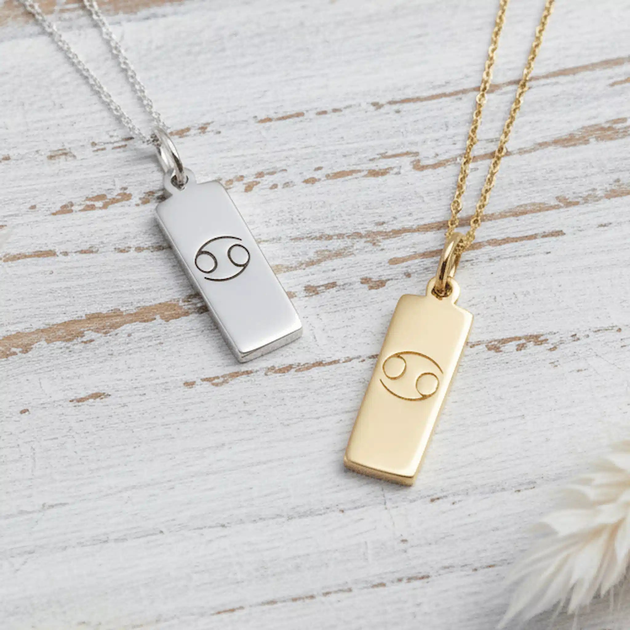 Two necklaces with rectangular pendants featuring cancer zodiac symbols on a wooden surface.