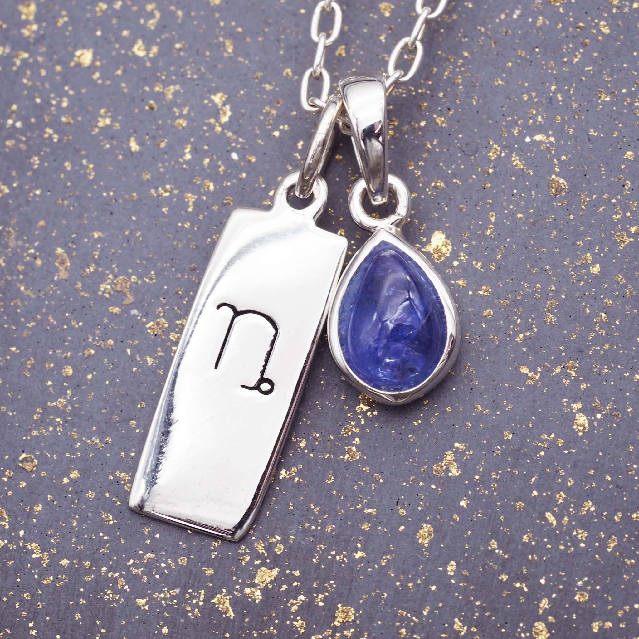 Silver necklace with capricorn zodiac charm and blue teardrop tanzanite pendant on a textured gray background