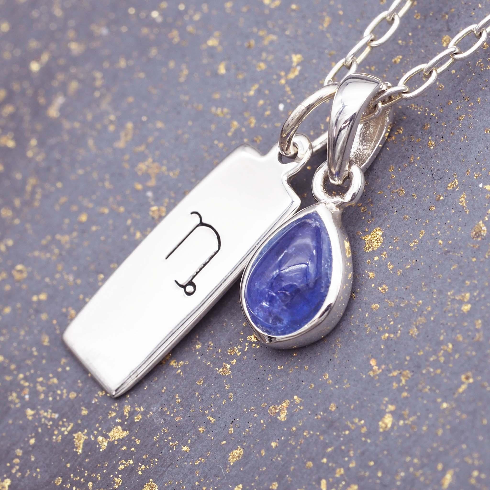 Silver necklace with a teardrop-shaped blue tanzanite gemstone pendant and a silver capricorn zodiac pendant on a textured gray background.