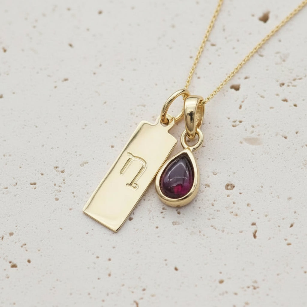 Gold necklace with Capricorn Zodiac symbol pendant and a teardrop-shaped natural garnet gemstone pendant on a light stone surface