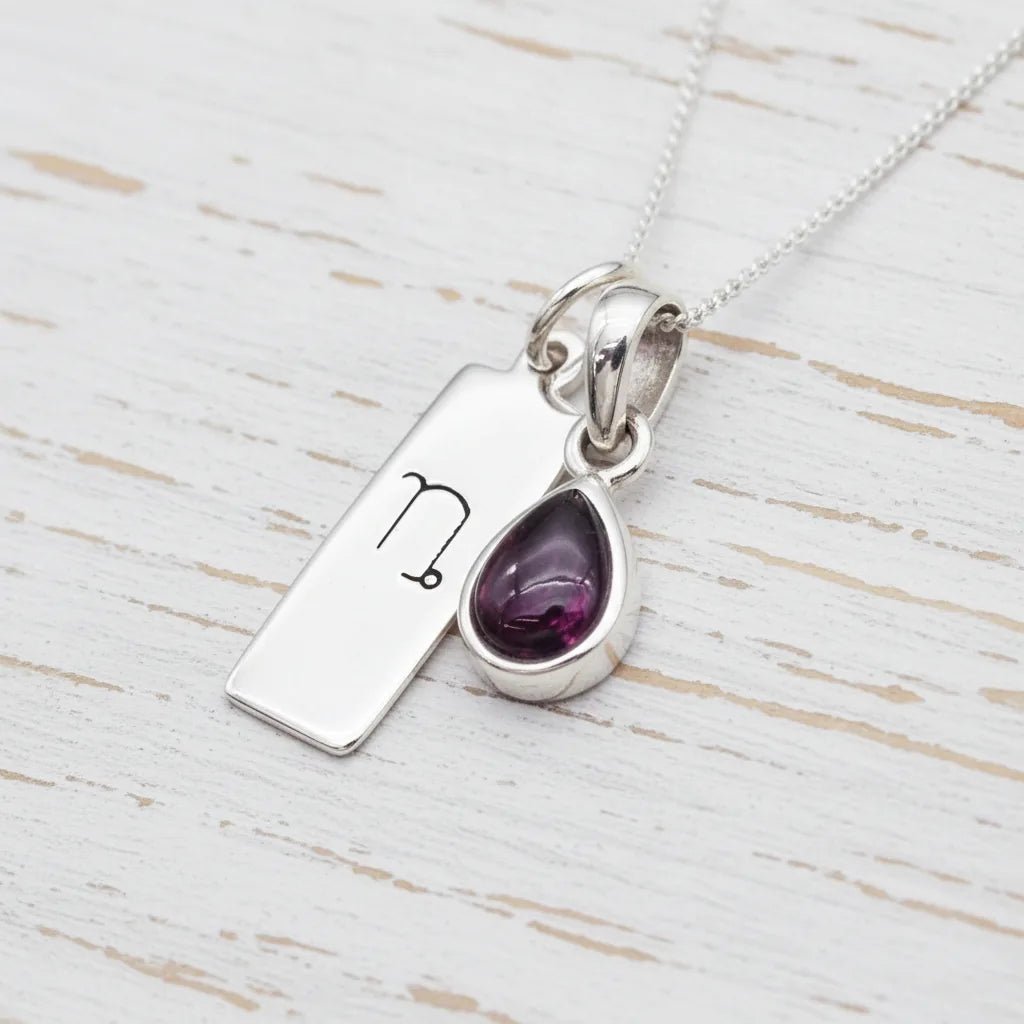 Silver necklace with a Capricorn Zodiac pendant together with a teardrop-shaped garnet gemstone pendant on a light wooden surface