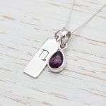 Silver necklace with a Capricorn Zodiac pendant together with a teardrop-shaped garnet gemstone pendant on a light wooden surface