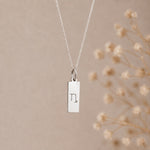capricorn zodiac necklace - A silver rectangular pendant necklace engraved with a minimalist capricorn star sign line glyph symbol, hanging from a delicate silver chain against a warm beige background