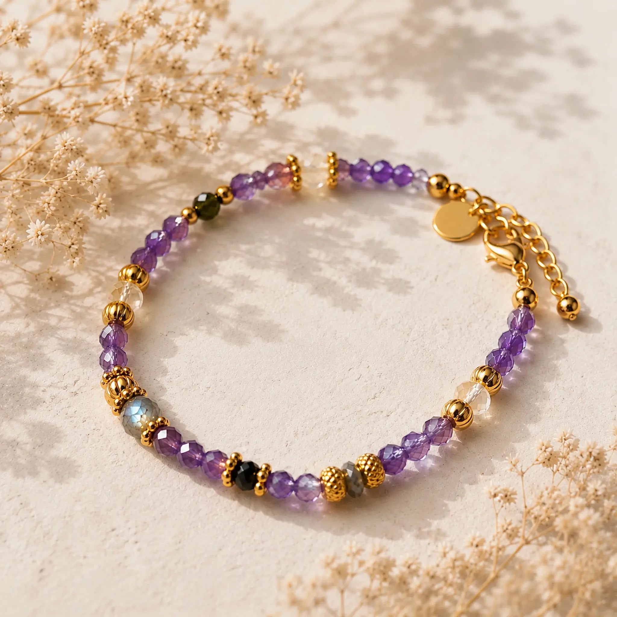 A delicate beaded bracelet with amethyst faceted beads, gold spacer beads, and a few dark green and iridescent blue-green accent beads, displayed on a textured beige surface with dried flowers.
