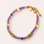 A beaded bracelet with faceted amethyst beads, textured gold spacer beads, a gray labradorite stone, and a small dark green bead, displayed on a light cream background.