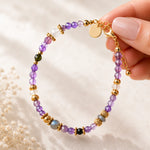 A delicate beaded bracelet with amethyst, citrine, aventurine, moonstone and labradorite faceted beads, accented by gold spacer beads and a gold clasp, held by a hand against a soft beige background.