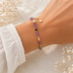 A delicate beaded bracelet with purple, gold, and white beads worn on a person's wrist, styled with white baby's breath flowers and a white ruffled sleeve.