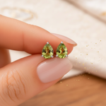 Hand holding a pair of faceted pear-shaped peridot stud earrings against a soft beige background