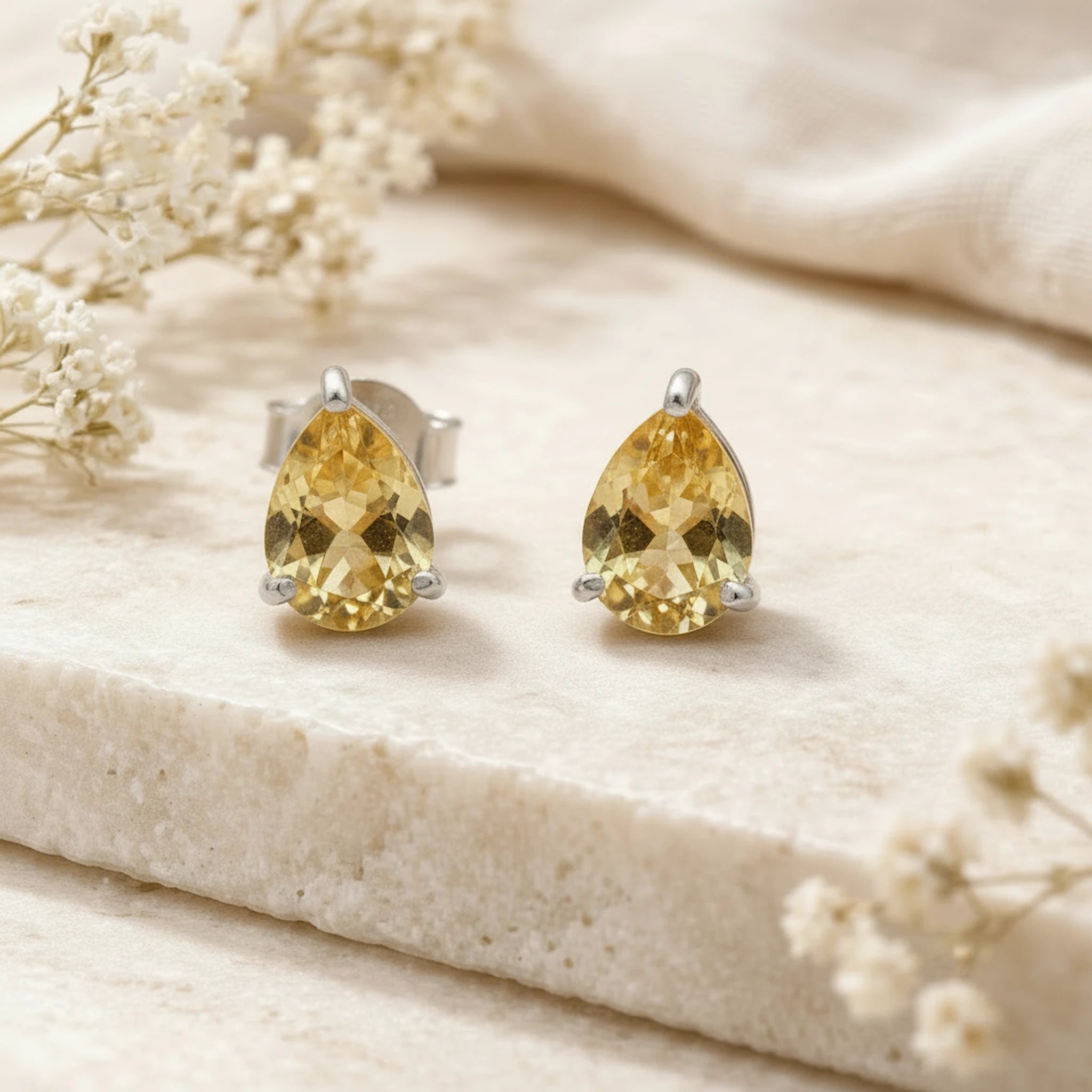 Pair of teardrop-shaped yellow citrine gemstone stud earrings on a beige surface with white flowers.
