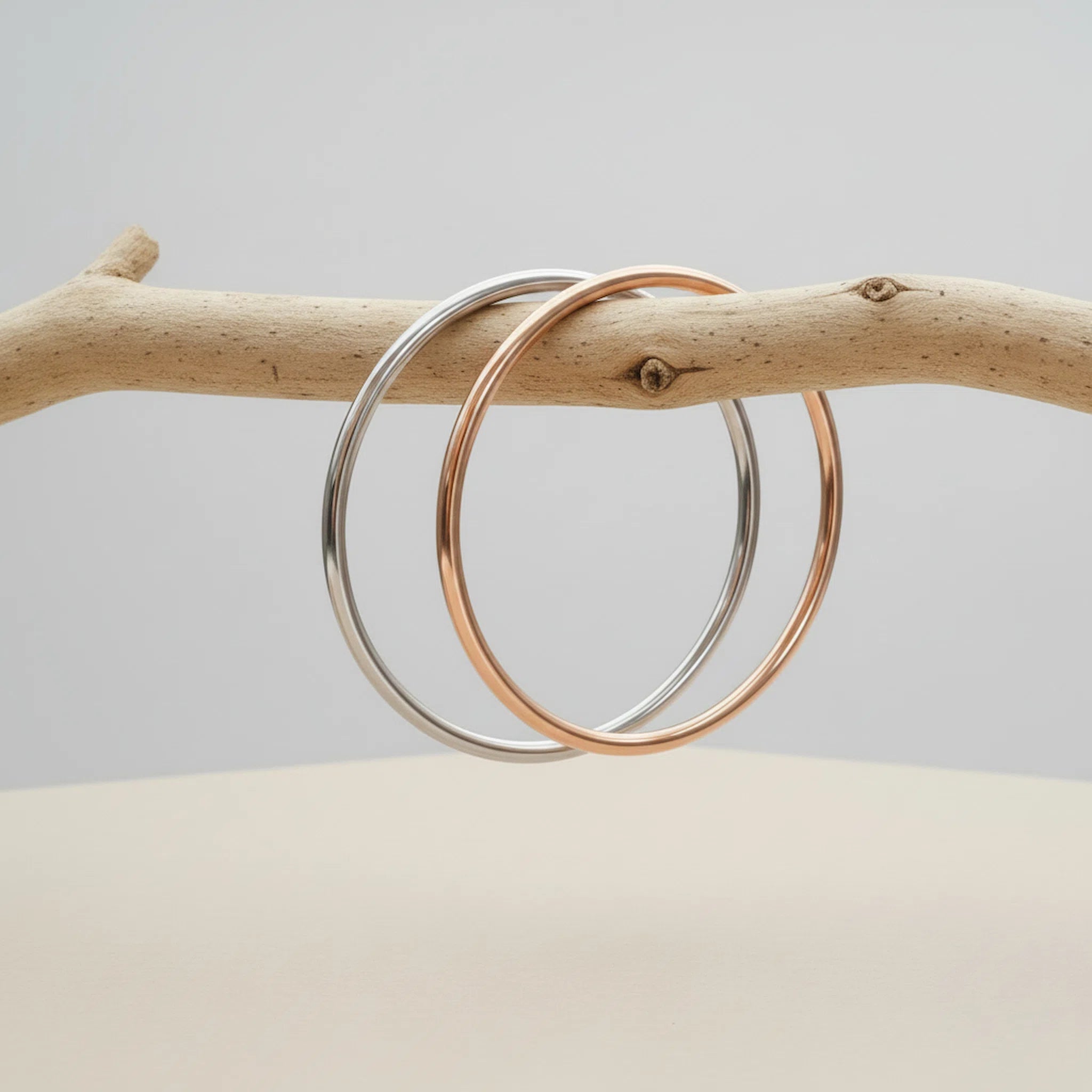 Two bangles, one silver and one rose gold, on a branch with a neutral background.