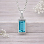 Silver waterproof necklace with a blue gemstone pendant on a wooden surface with white flowers.