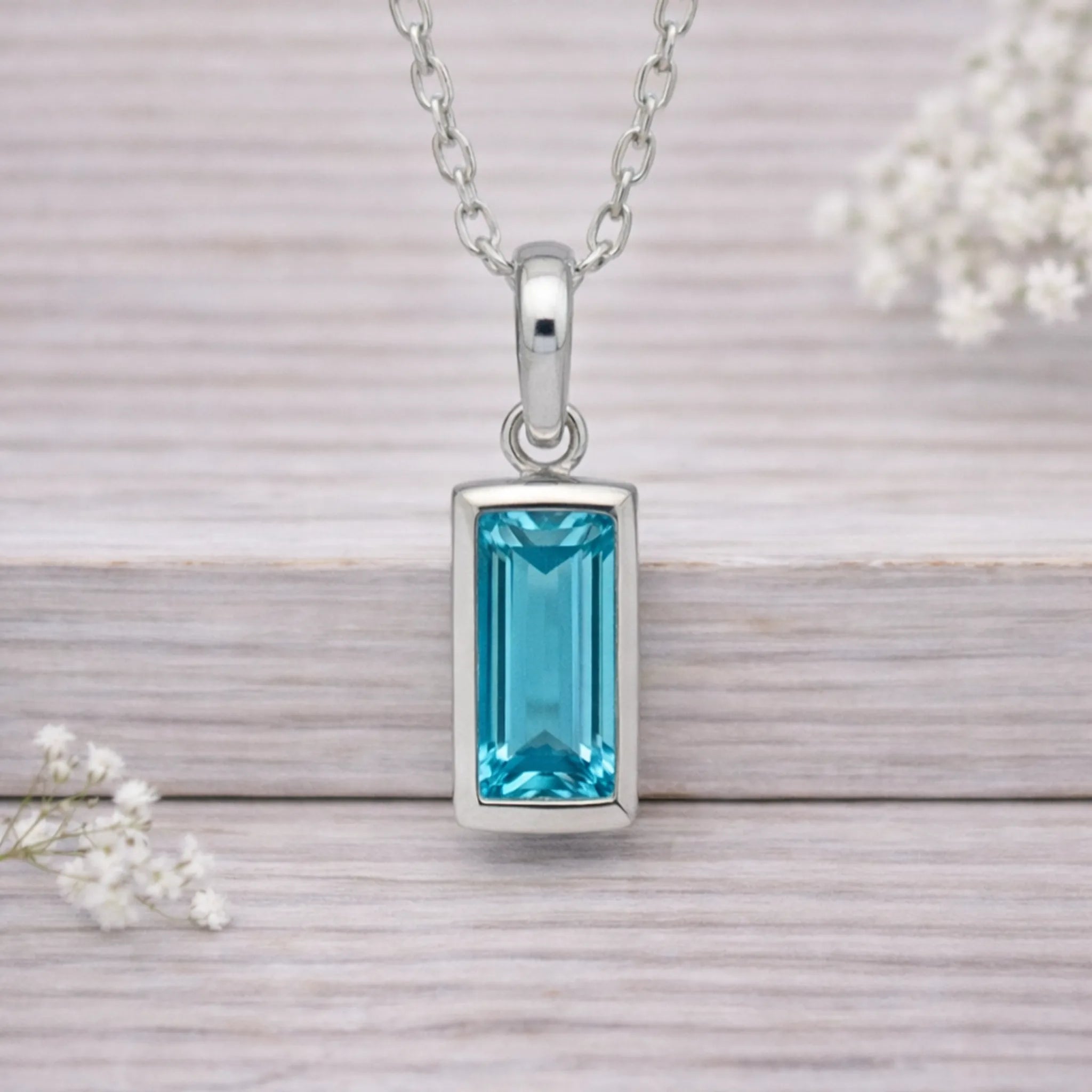 Silver waterproof necklace with a blue gemstone pendant on a wooden surface with white flowers.