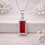 Silver waterproof necklace with a red gemstone pendant on a wooden surface with white flowers.