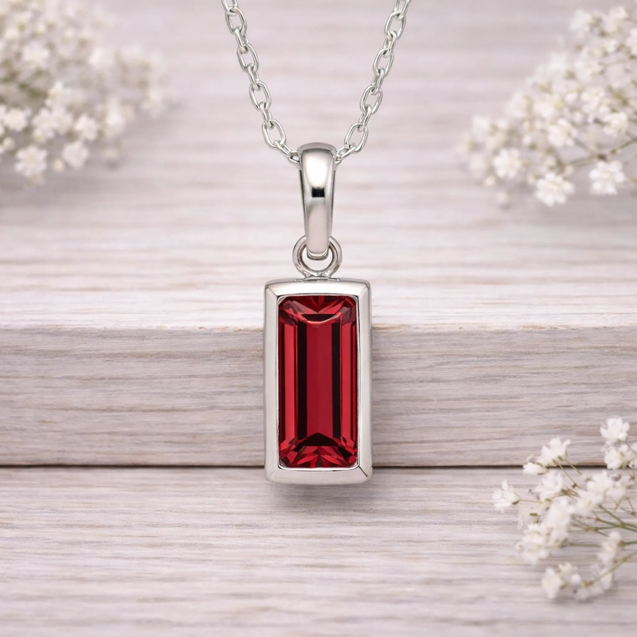 Silver necklace with a red gemstone pendant on a wooden surface with white flowers.