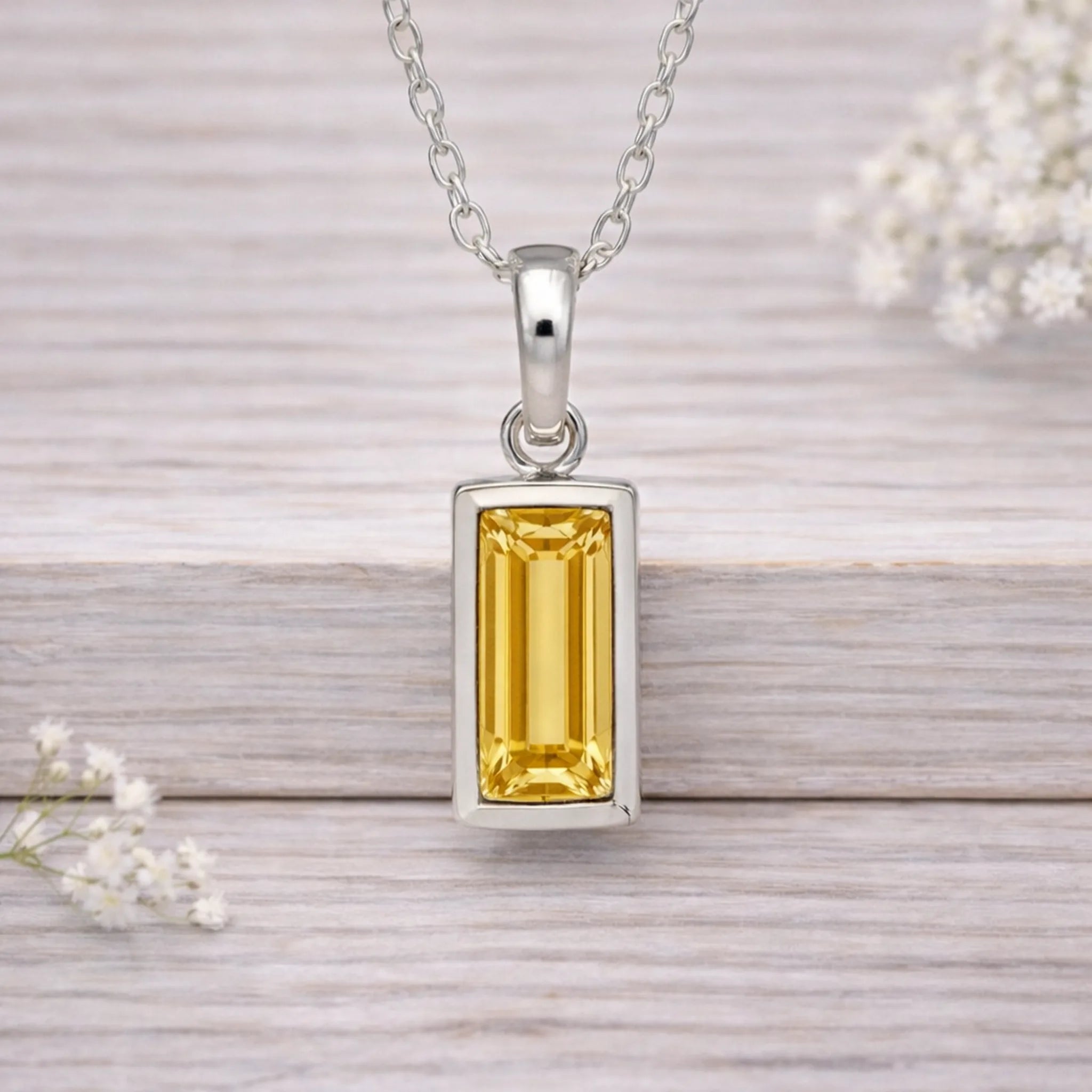 Silver waterproof necklace pendant with a yellow gemstone on a wooden surface with white flowers.