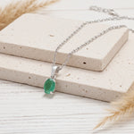 A silver chain necklace with an oval green gemstone pendant, displayed on stacked beige stone blocks with dried grass accents on a light wood surface.