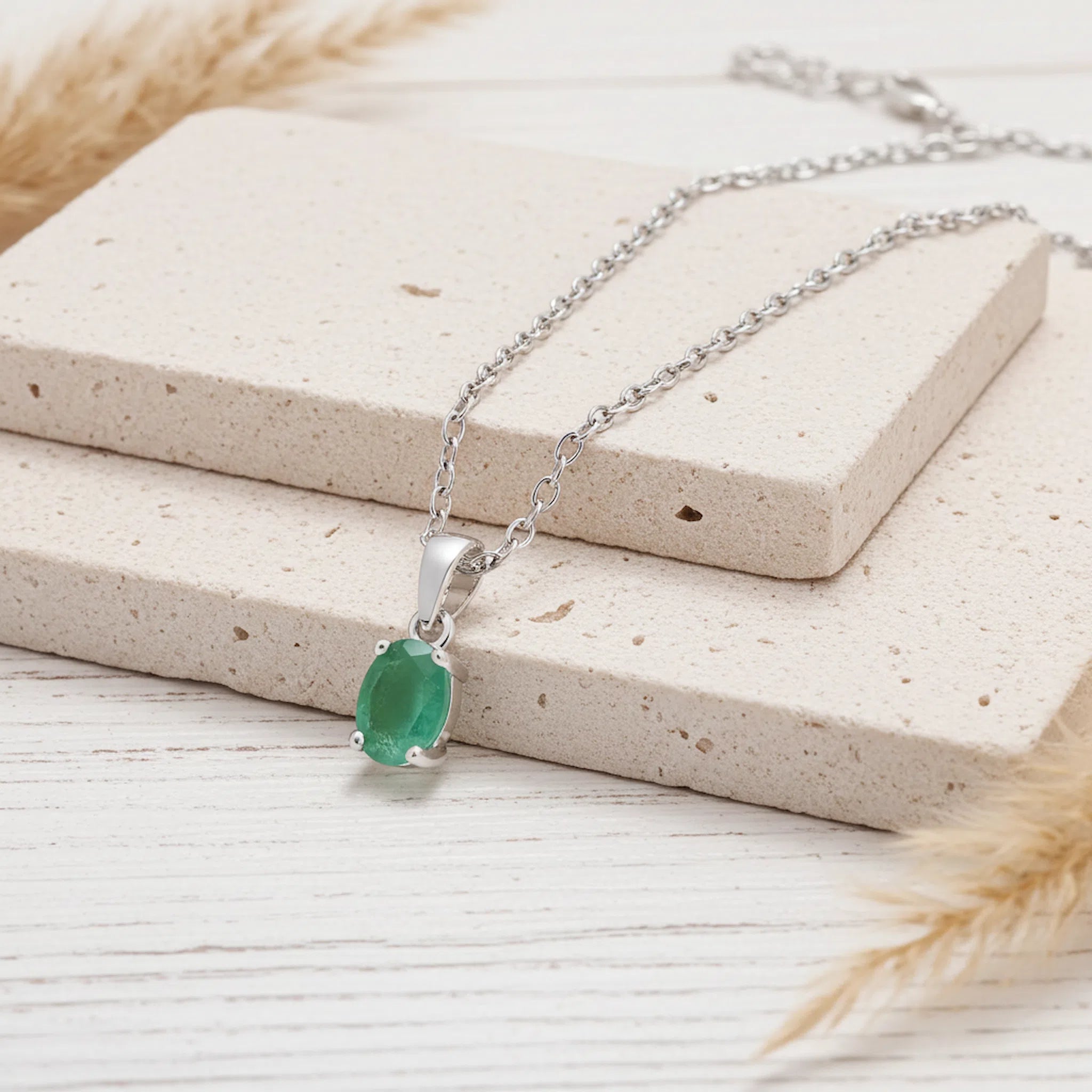 A silver chain necklace with an oval green gemstone pendant, displayed on stacked beige stone blocks with dried grass accents on a light wood surface.