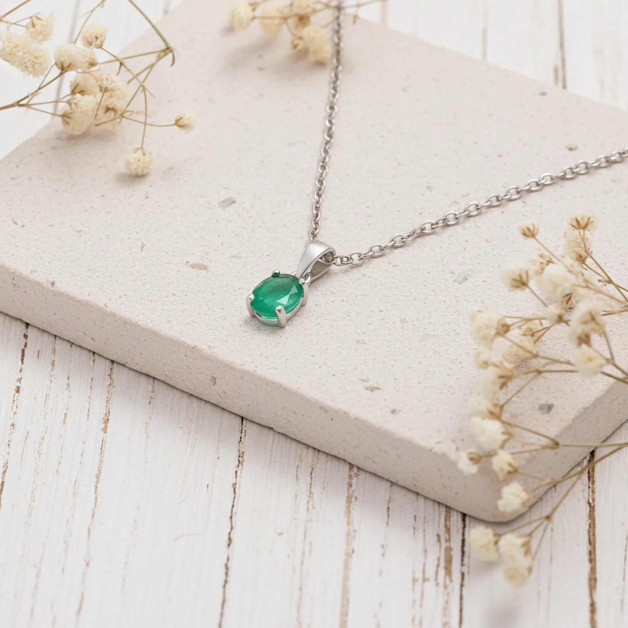 A silver pendant necklace with a green oval-cut gemstone, displayed on a light beige stone slab with dried baby's breath flowers on a white wooden surface.