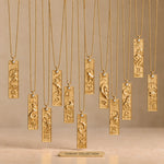 Cosmic Collection of twelve gold zodiac bar necklaces on fine ball chains, arranged at staggered heights over a warm beige background and smooth matte base