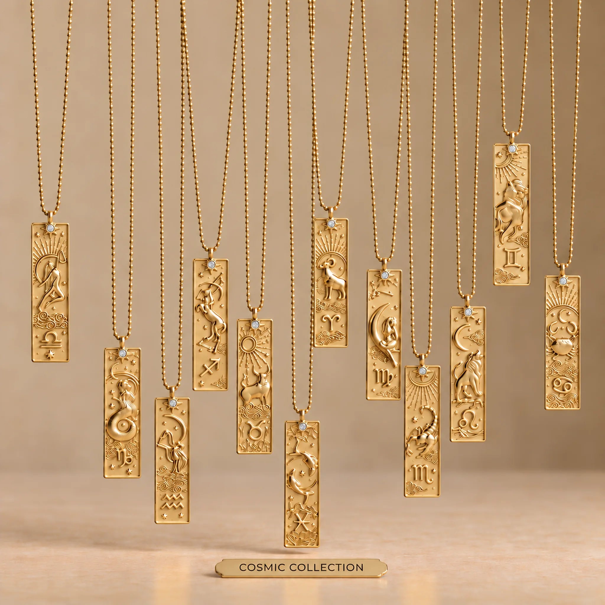 Cosmic Collection of twelve gold zodiac bar necklaces on fine ball chains, arranged at staggered heights over a warm beige background and smooth matte base
