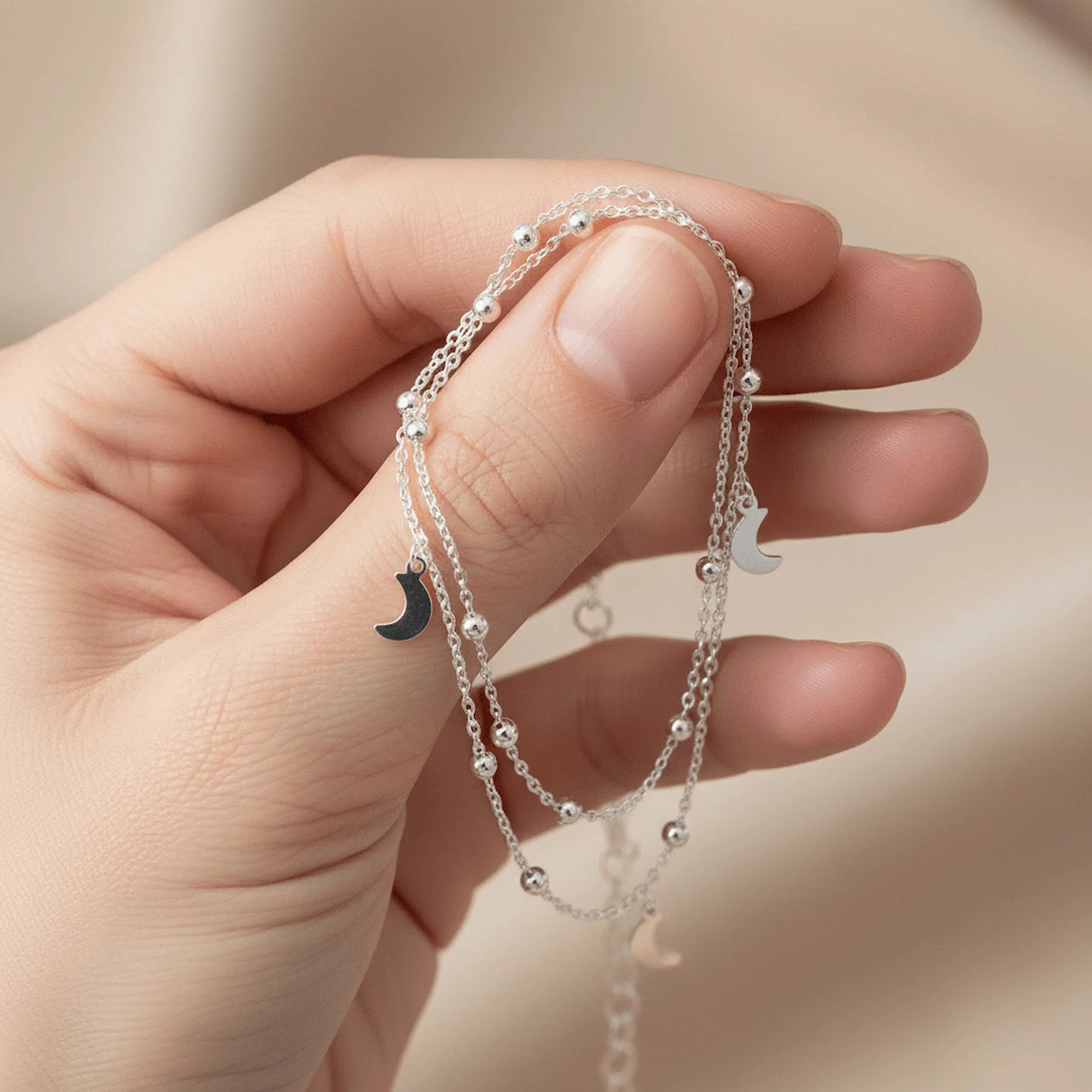 Hand holding silver anklet with moon pendants on a neutral background