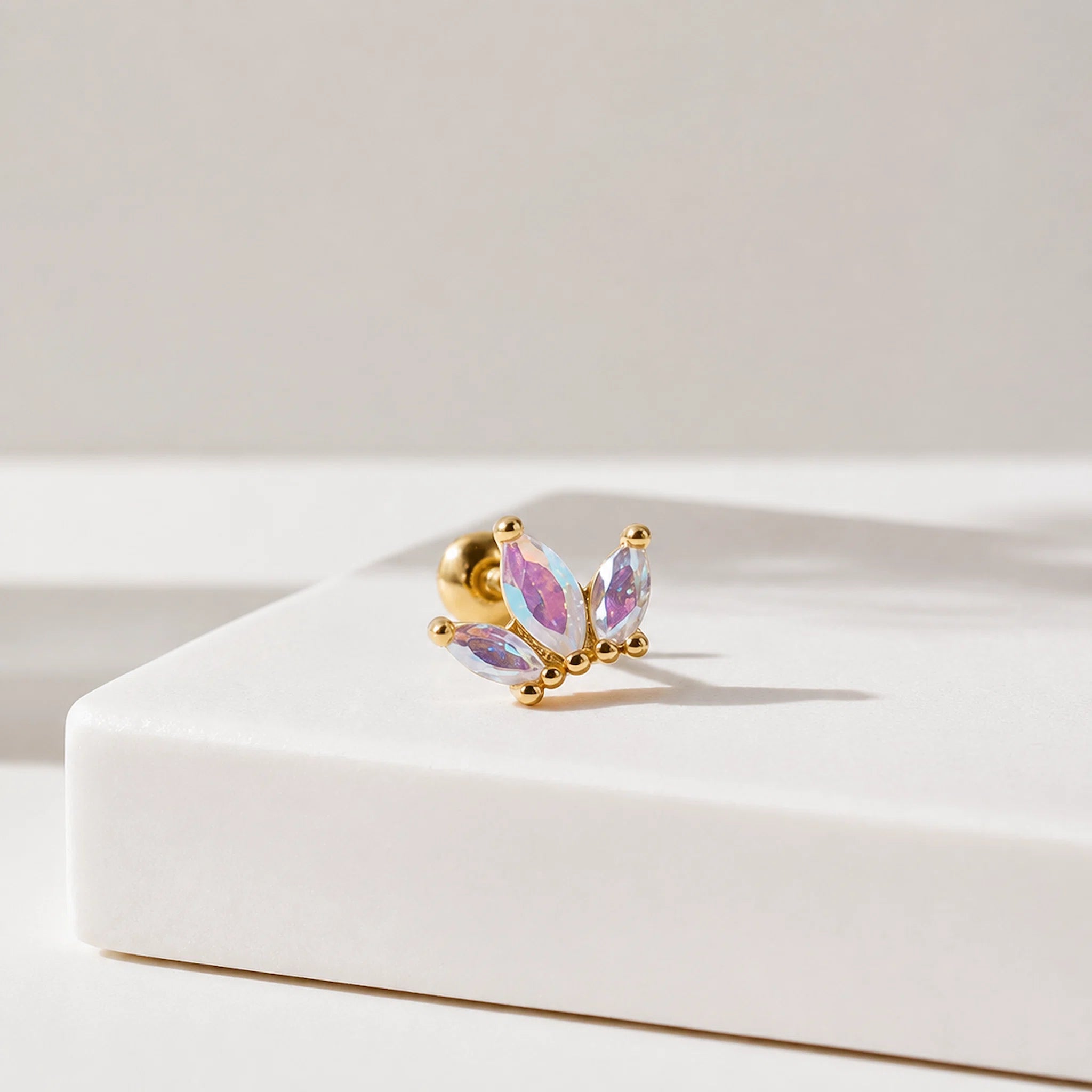 A gold post piercing stud featuring three marquise-cut iridescent crystals, displayed on a clean white geometric block against a neutral light gray background.