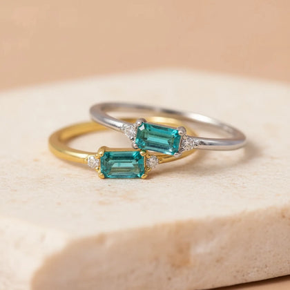 Two dainty blue green tourmaline rings in sterling silver and gold plated sterling silver with rectangular faceted centre stones and round cubic zirconia side accents on a beige stone surface