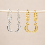Two pairs of hoop earrings with crescent moon charms hang from a white rod against a light beige background. The left pair is silver, and the right pair is gold.